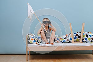 Little boy is sitting on unmade bed with white flag, looking through binoculars