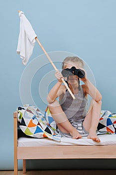 Little boy is sitting on unmade bed with white flag, looking through binoculars