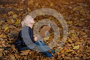 Little boy is sitting in wild forest in autumn leaves with book in hands. Children`s books about miracles and magic