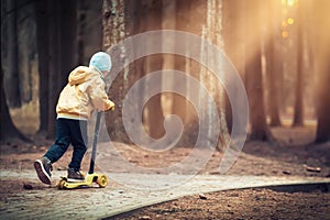 Little boy skates on scooter in evening park at sunset under light of lanterns. kid is riding scooter along path in dark forest.