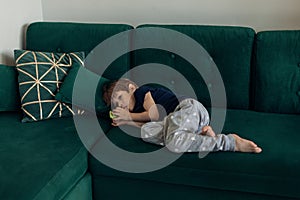 Little boy in sleepwear lying on big green modern sofa in living room and sucking finger. Bad habbit and manners of photo