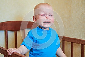 A little boy is standing in the crib, crying and calling mother