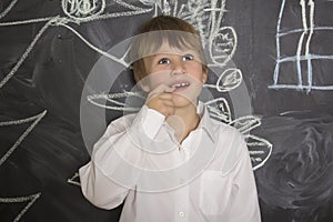 Little boy standing near the blackboard for drawing. Fingers holding in the mouth.