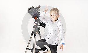 A little boy stands at a telescope and points his finger upwards.