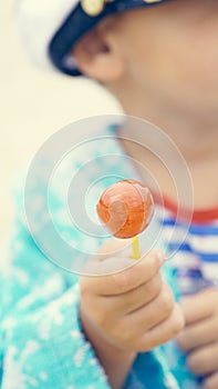 Little boy in a striped t-shirt and captain`s cap sitting on the beach with candy in mouth