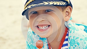 Little boy in a striped t-shirt and captain`s cap sitting on the beach with candy in mouth