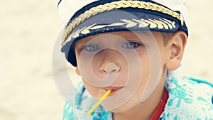 Little boy in a striped t-shirt and captain`s cap sitting on the beach with candy in mouth