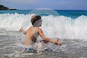 little boy in swimming goggles sitting on pebble on beach and enjoys surging waves with white foam