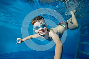 Little boy swims underwater in the pool, smiling, blowing bubbles and looking at me. The view from under the water. Close-up