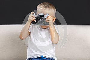 A little boy takes pictures. A child in a white T-shirt and jeans sits on a white sofa on a black background and holds a camera in