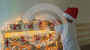 Little boy takes a present from an advent calendar hanging on a bed that is lightened with Christmas lights. Getting