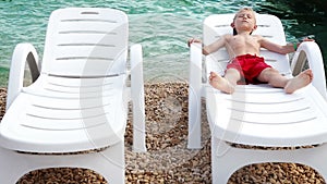 Little boy tans on the sea coastline in white deck chair