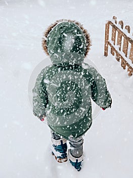 A little boy of two years is standing with his back in a green jacket with a hood, gray pants and blue boots, and in the