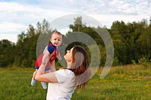 A little boy up to a year old in the arms of his mother plays, laughs and smiles against the background of a natural green landsca