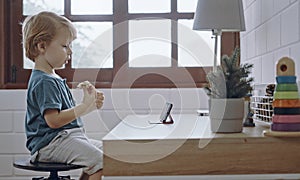 Little boy using smartphone, looking at screen, curious child holding phone in hands, sitting on chair at home alone