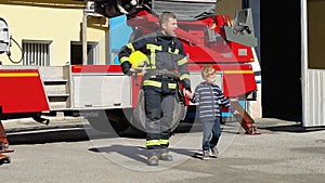 Little boy walk with firefighter in protective uniform in fire station