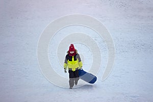 A little boy in warm bright outerwear carries a sled