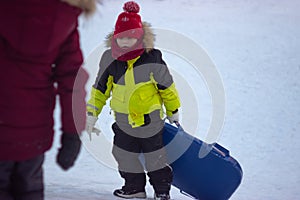 A little boy in a warm yellow jacket carries a sled up the hill to slide down