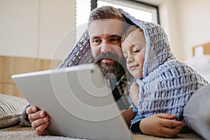 Little boy watching cartoon movie on tablet with father, lying under blanket on floor in kids room. Dad explaining