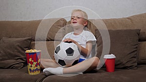 Little boy is watching a football match on TV sitting on the couch with popcorn.