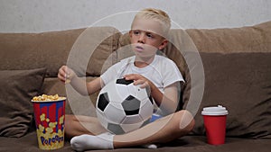 A little boy is watching a football match on TV sitting on couch with popcorn.