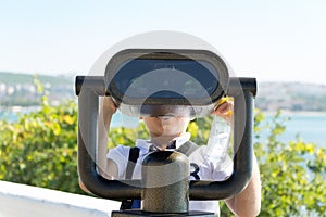 Little boy watching through stationary binocular from promenade at sunny day in vacation. Facing to the viewer