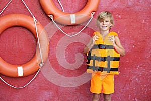 Little boy wearing orange life vest near red wall with safety rings