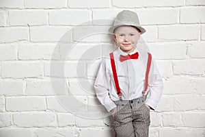 Little boy wearing a red bow tie, suspenders and white shirtand against a white brick wall