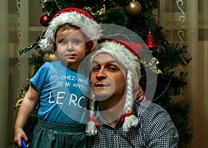 A little boy wearing Santa Claus hat and his uncle wearing Snow Maiden hat sitting on background of New Year tree.