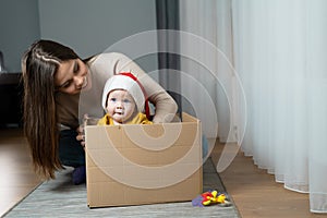 A little boy wearing a Santa Claus hat sits in a box on the floor next to his Caucasian mother. Happy family on