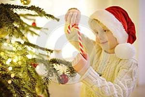 Little boy wearing Santa hat ready for celebrate Christmas. Cute child decorating the Christmas tree with big sweet candy. Baby
