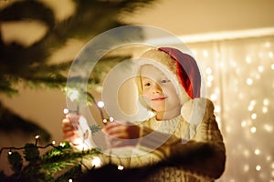 Little boy wearing Santa hat ready for celebrate Christmas. Cute child decorating the Christmas tree with garland. Baby hopes of