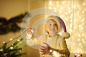 Little boy wearing Santa hat ready for celebrate Christmas. Cute child decorating the Christmas tree with garland. Baby hopes of