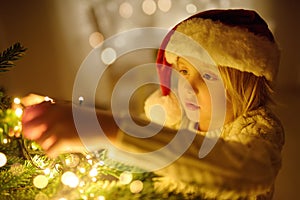 Little boy wearing Santa hat ready for celebrate Christmas. Cute child decorating the Christmas tree with garland. Baby hopes of