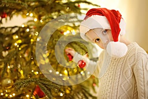 Little boy wearing Santa hat ready for celebrate Christmas. Cute child decorating the Christmas tree with glass toy. Baby hopes of