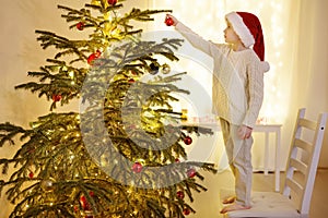 Little boy wearing Santa hat ready for celebrate Christmas. Cute child decorating a large Christmas tree with ornate toys. Baby