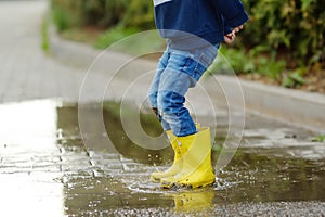 Little boy wearing yellow rubber boots jumping and having fun in puddle of water on rainy summer day in small town. Outdoors games