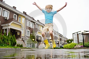 Little boy wearing yellow rubber boots jumping in puddle of water on rainy summer day in small town. Child having fun