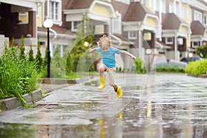Little boy wearing yellow rubber boots jumping in puddle of water on rainy summer day in small town. Child having fun