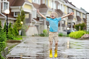 Little boy wearing yellow rubber boots jumping in puddle of water on rainy summer day in small town. Child having fun