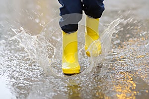 Little boy wearing yellow rubber boots jumping in puddle of water on rainy summer day in small town. Child having fun