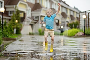 Little boy wearing yellow rubber boots jumping in puddle of water on rainy summer day in small town. Child having fun