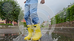 Little boy wearing yellow rubber boots jumping in puddle of water on rainy summer day in small town. Child having fun.