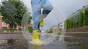 Little boy wearing yellow rubber boots jumping in puddle of water on rainy summer day in small town. Child having fun.