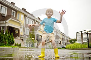 Little boy wearing yellow rubber boots jumping in puddle of water on rainy summer day in small town. Child having fun. Outdoors