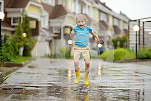 Little boy wearing yellow rubber boots jumping in puddle of water on rainy summer day in small town. Child having fun. Outdoors