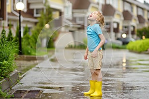 Little boy wearing yellow rubber boots walking on rainy summer day in small town. Child having fun. Kid catches raindrops with