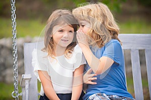 Little boy whispers to lovely girl in ear. Brother and sister playing in spring park outdoors. Little boy and girl kids