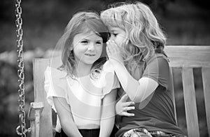 Little boy whispers to lovely girl in ear. Brother and sister playing in spring park outdoors. Little boy and girl kids