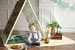 Little boy in a white shirt, jeans and bow tie is sitting in a tent in his room decorated in the style of happy Easter